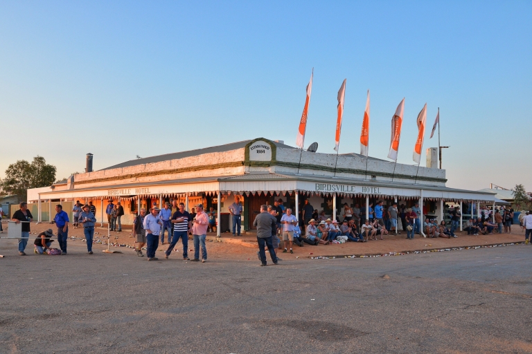 A historic pub with a wrap-around awning decorated with flags and a crowd of people gathered around outside sitting, drinking and talking at Birdsville Hotel, Birdsville, Queensland © Tourism and Events Queensland A historic pub with a wrap-around awning decorated with flags and a crowd of people gathered around outside sitting, drinking and talking at Birdsville Hotel, Birdsville, Queensland © Tourism and Events Queensland