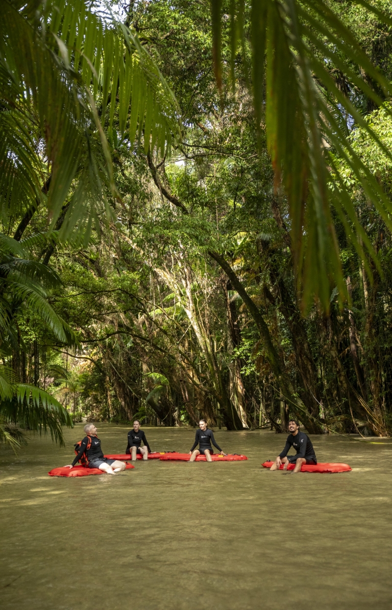 A group of people sitting on inflatable red boards, floating on a calm river beneath a lush green rainforest canopy with Back Country Bliss Adventures, Daintree Rainforest, Queensland © Tourism and Events Queensland A group of people sitting on inflatable red boards, floating on a calm river beneath a lush green rainforest canopy with Back Country Bliss Adventures, Daintree Rainforest, Queensland © Tourism and Events Queensland