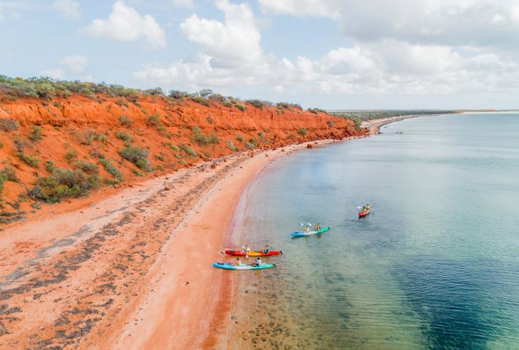 Aerial view over a group of people kayaking in clear blue water beside bright orange cliffs during a tour with Wula Gura Nyinda Eco Cultural Adventures, Coral Coast, Western Australia © Tourism Western Australia