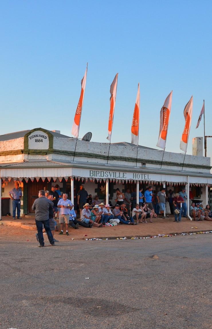A historic pub with a wrap-around awning decorated with flags and a crowd of people gathered around outside sitting, drinking and talking at Birdsville Hotel, Birdsville, Queensland © Tourism and Events Queensland