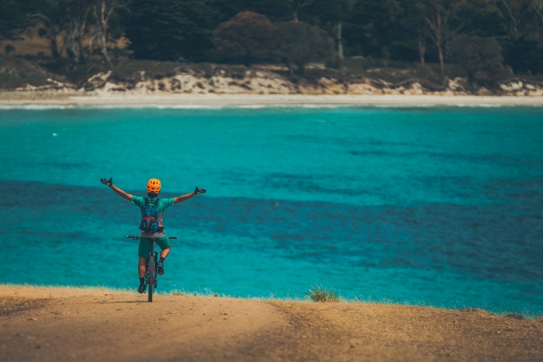 Une personne sur un VTT, bras levés en l'air, surplombant l'océan limpide dans le Maria Island National Park, Tasmanie © Matt Staggs Une personne sur un VTT, bras levés en l'air, surplombant l'océan limpide dans le Maria Island National Park, Tasmanie © Matt Staggs