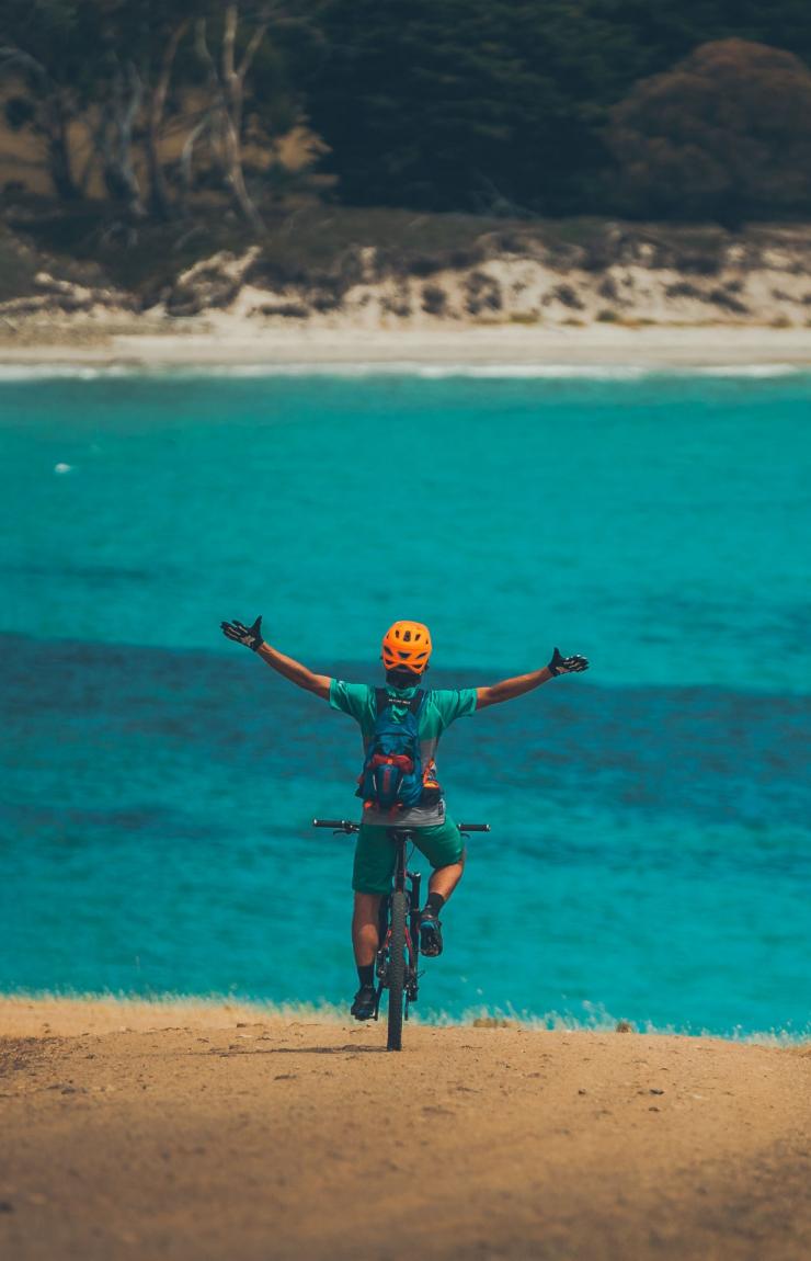 Une personne sur un VTT, bras levés en l'air, surplombant l'océan limpide dans le Maria Island National Park, Tasmanie © Matt Staggs