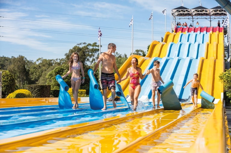 Famille souriante marchant au pied d'un toboggan jaune, bleu et rouge au parc aquatique Wet'n'Wild de la Gold Coast, Queensland © Destination Gold Coast Famille souriante marchant au pied d'un toboggan jaune, bleu et rouge au parc aquatique Wet'n'Wild de la Gold Coast, Queensland © Destination Gold Coast