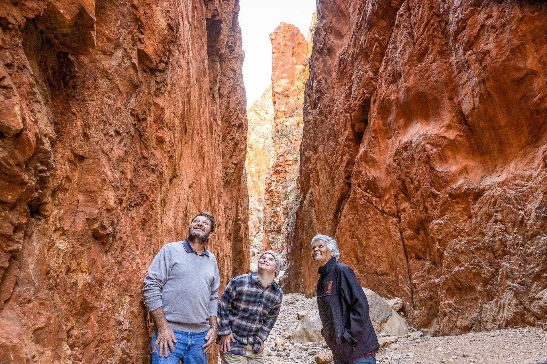 Seorang pria dengan neurodivergensi didampingi oleh pria lain dan seorang pemandu tur di Standley Chasm, West MacDonnell Ranges, Northern Territory © Tourism NT/Helen Orr