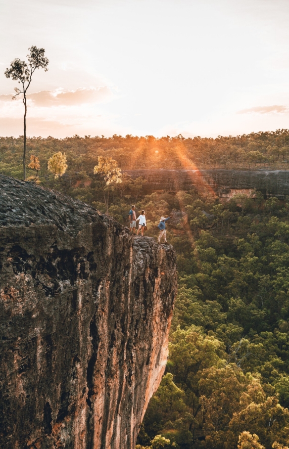 Seorang pemandu wisata Aborigin dan dua tamu berdiri di atas singkapan berbatu yang menghadap padang semak hijau dalam tur bersama Jarramali Rock Art Tours, Tropical North Queensland, Queensland © Tourism Tropical North Queensland 