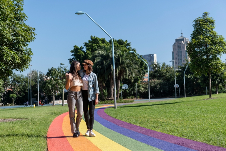 Pasangan sedang berjalan berangkulan menelusuri jalur pelangi di antara rumput hijau dan pepohonan di Prince Alfred Park, Surry Hills, New South Wales © Destination NSW