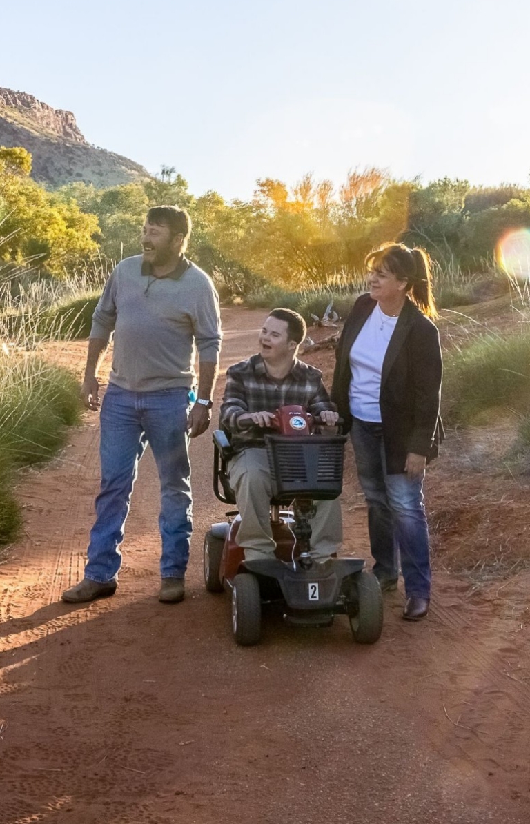 Uomo seduto sulla carrozzina di fianco ad un altro uomo e ad una donna ammirano la vegetazione lussureggiante e la sabbia rossa di Alice Springs, nell'Alice Spring Desert Park, Northern Territory. Uomo seduto sulla carrozzina di fianco ad un altro uomo e ad una donna ammirano la vegetazione lussureggiante e la sabbia rossa di Alice Springs, nell'Alice Spring Desert Park, Northern Territory.