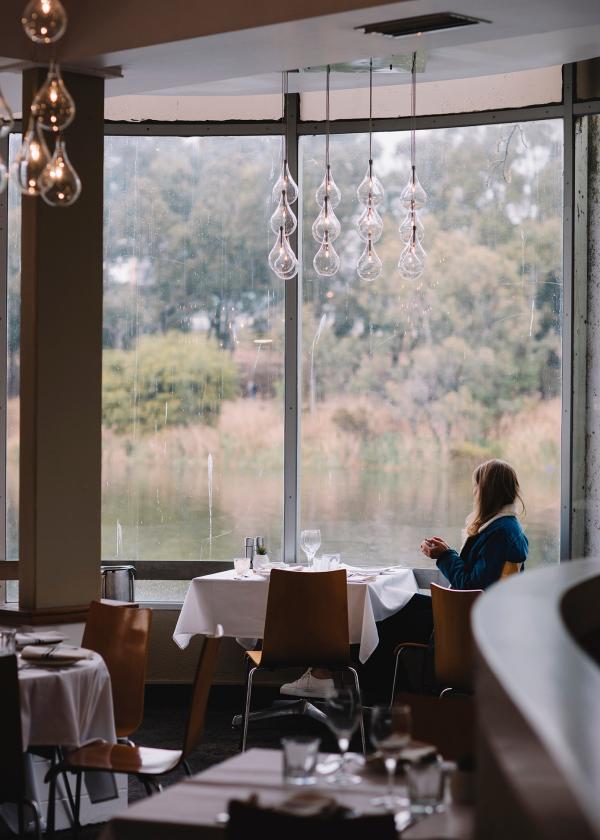 Persona seduta in un ristorante accanto a una finestra a parete intera che ammira la vista su un tranquillo corso d'acqua al River Café, North Adelaide, South Australia © Tourism Australia