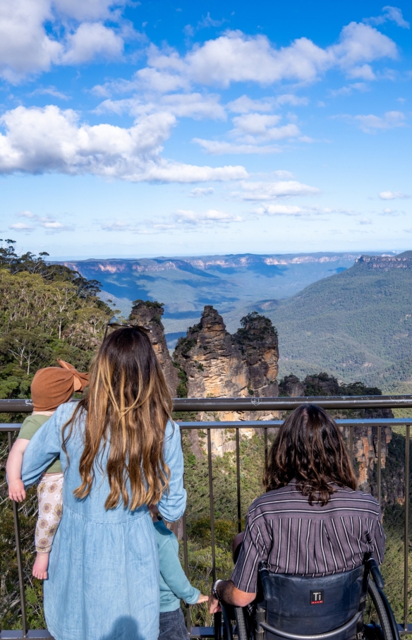 Uomo su una sedia a rotelle con la sua famiglia che ammirano le Three Sisters nel Blue Mountains National Park, New South Wales © Tourism Australia