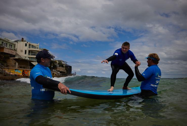 Uomo con neurodivergenza che si alza su una tavola da surf con l'aiuto del Let's Go Surfing, Bondi Beach, Sydney, New South Wales © Tourism Australia