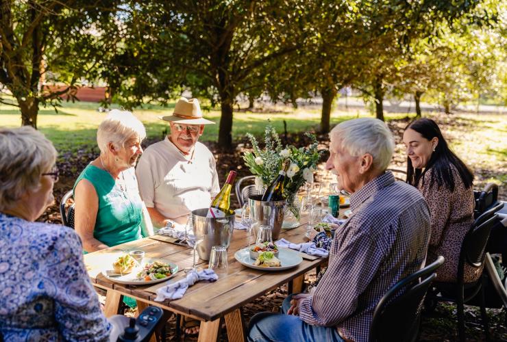 Amici seduti a mangiare piatti a base di macadamia tra gli alberi di macadamia al Macadamias Australia, Bundaberg, Queensland © Tourism Australia