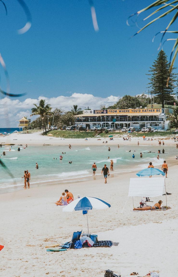Persone sdraiate sulla sabbia bianca sotto ombrelloni colorati e altre persone che nuotano tra le placide onde e nelle acque cristalline di Greenmount Beach a Coolangatta, Queensland © Tourism and Events Queensland Persone sdraiate sulla sabbia bianca sotto ombrelloni colorati e altre persone che nuotano tra le placide onde e nelle acque cristalline di Greenmount Beach a Coolangatta, Queensland © Tourism and Events Queensland