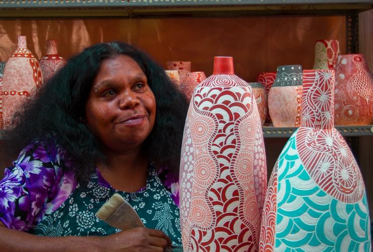 ノーザンテリトリー、ウルル、ロンギチュード131、エルナベラ・アートにて、陶芸品の横に腰掛けるアボリジナル・アーティスト © Longitude 131