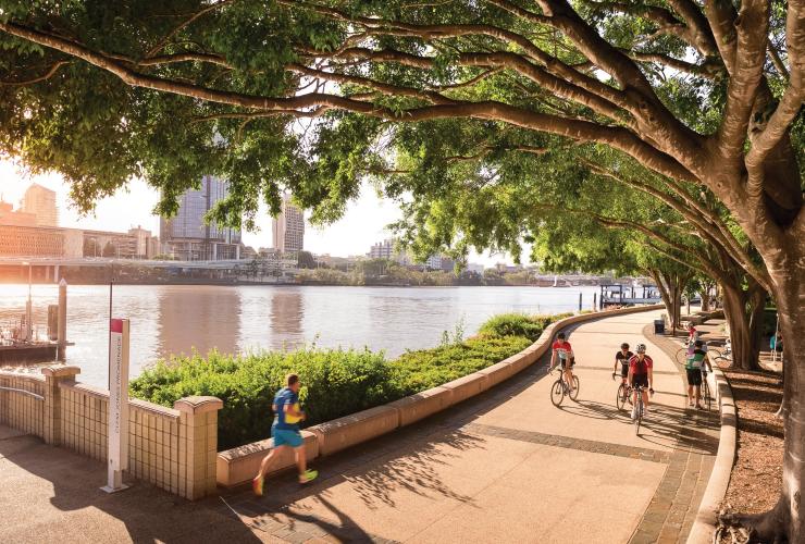 Eine Gruppe von Radfahrern fährt auf einem begrünten Weg entlang eines Flusses mit hohen Gebäuden im Hintergrund, River Walk, South Bank, Brisbane, Queensland © Fullframe Photographics