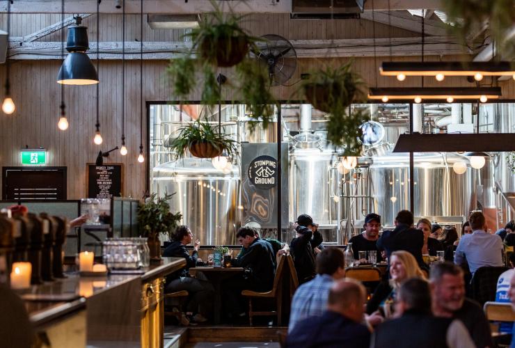 Ein gut besuchtes Lokal, in dem Menschen lächelnd Bier an Tischen trinken, während man durch große Fenster die Brauereiausrüstung der Stomping Ground Brewery Co. sehen kann, Collingwood, Victoria © Stomping Ground Brewery Co.
