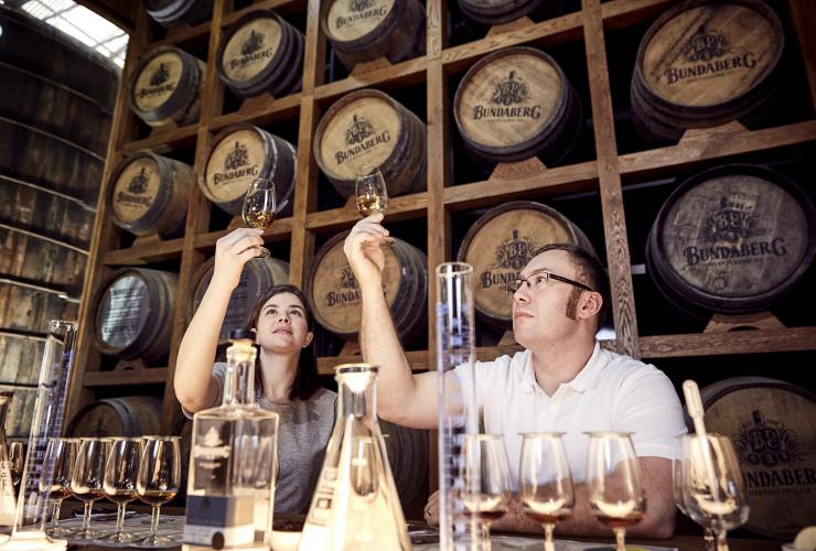 Ein Mann und eine Frau schauen in mit Rum gefüllte Gläser, die sie während einer Blend Your Own Rum Experience in der Bundaberg Rum Distillery gegen das Licht halten, Bundaberg, Queensland © Bundaberg Rum