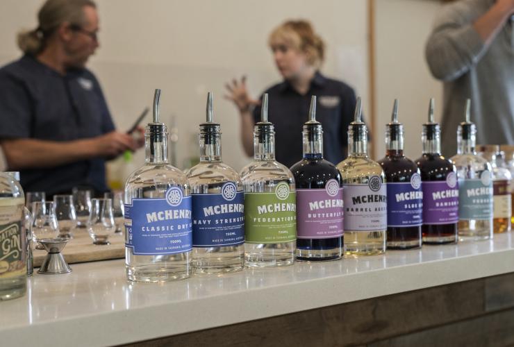 Spirituosen mit verschiedenfarbigen Etiketten sind auf einem Tisch aufgereiht, während sich im Hintergrund Gäste unterhalten, McHenry Distillery, Tasman Peninsula, Tasmanien © Tourism Australia