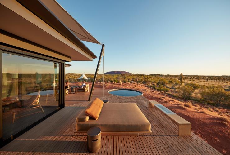 Außenaufnahme der Unterkunft des Longitude 131 in Yulara mit zwei Personen, die auf einer privaten Terrasse mit Pool in Clubsesseln sitzen und die ungestörte Aussicht auf den Uluru im Hintergrund genießen, Northern Territory © Baillie Lodges