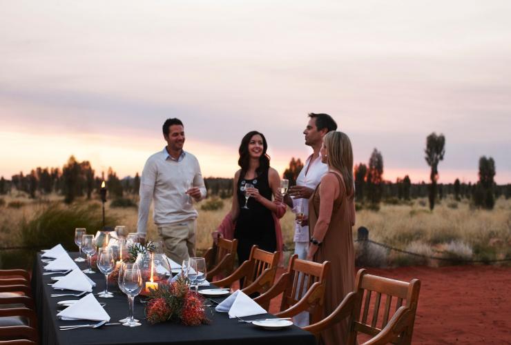 Vier Personen sitzen mit Weingläsern in der Hand an einem Tisch, der mit Kerzen und Blumen gedeckt ist, unter freiem Himmel in der Wüste im Table 131, Longitude 131, Uluru-Kata Tjuta National Park, Northern Territory ©Luxury Lodges of Australia