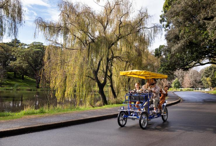 Une famille conduisant une rosalie recouverte d'un parasol le long d'un étang, près de la végétation des sentiers Centennial Park Cycles, Centennial Park, Nouvelle-Galles du Sud © Destination NSW