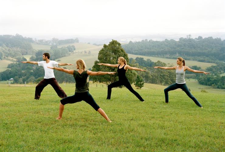Un groupe de personne exécutant une posture de yoga, les bras tendus, sur une colline verdoyante surplombant l'arrière-pays de Byron Bay à Gaia Retreat & Spa, Nouvelle-Galles du Sud © Tourism Australia