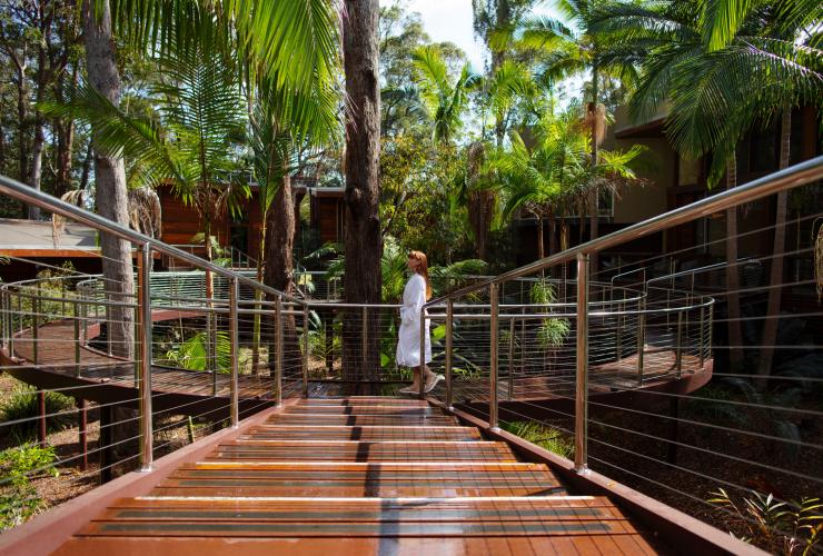 Une femme portant un peignoir blanc marchant sur une passerelle en bois suspendue entourée d'arbres, devant un bâtiment en bois au Gwinganna Lifestyle Retreat, Gold Coast, Queensland © Destination Gold Coast