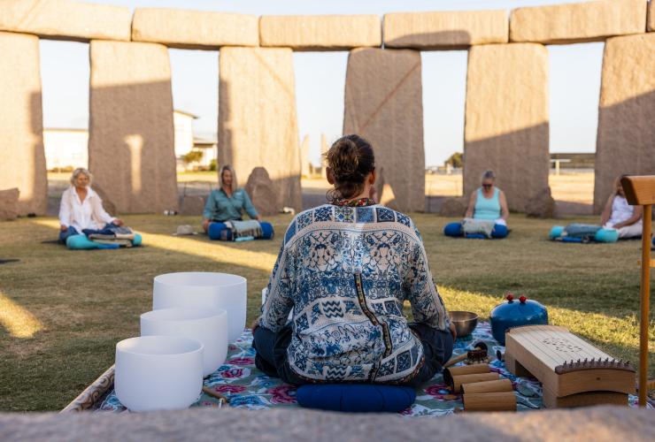 Une professeure de yoga de dos, assise à côté de plusieurs instruments, devant un groupe de personnes assises séparément réalisant des postures de yoga avec Untamed Escapes, Esperance, Australie Occidentale © Tourism Australia