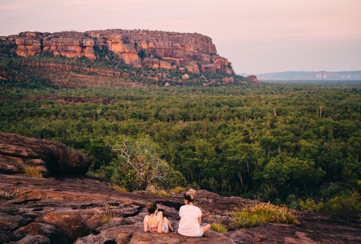 Dua orang duduk di atas tebing berbatu menghadap ke bentangan tanaman rimbun di Burrungkuy (Nourlangie) Rock, Kakadu National Park, NT © Tourism NT/Jewels Lynch