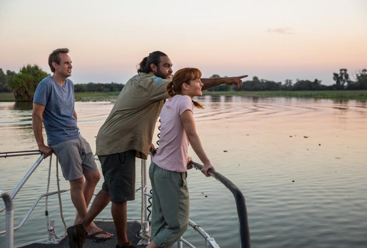 Dua orang berdiri bersama pemandu Aborigin sambil menunjuk ke seberang kolam billabong datar yang memantulkan semburat warna pastel senja saat pesiar Yellow Water Billabong, Kakadu Tourism, Northern Territory © Tourism Australia