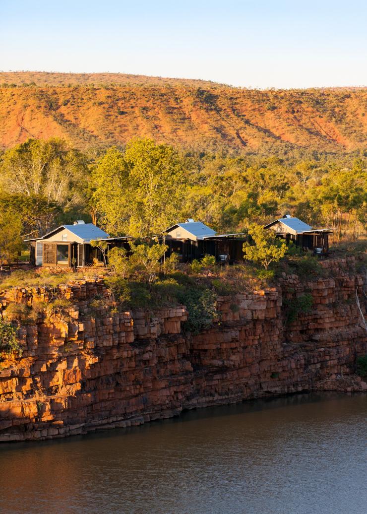 Akomodasi El Questro Homestead yang dikelilingi pepohonan hijau di tepi tebing batu merah yang mengarah ke sungai di El Questro Wilderness Park, Western Australia © Timothy Burgess