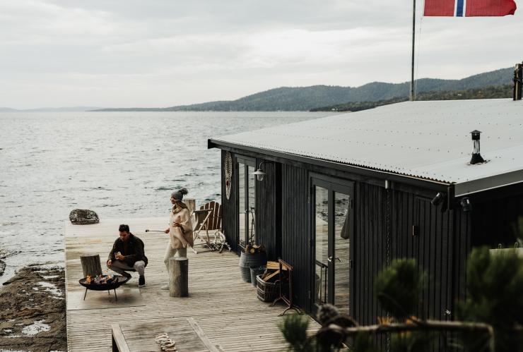 Dua orang berkumpul di sekeliling api unggun outdoor di balkon di samping pondok penginapan yang lebih tinggi dari permukaan samudra di Satellite Island, Tasmania © Adam Gibson