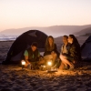 Sekelompok teman duduk mengelilingi cahaya di pantai saat matahari terbenam dengan tenda di belakang mereka, Tunkalilla Beach, Fleurieu Peninsula, South Australia © South Australian Tourism Commission/Peter Fisher