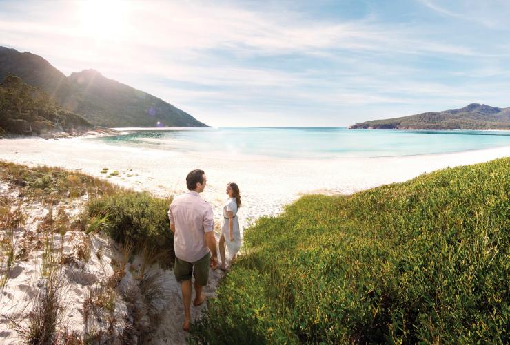 Pasangan berjalan melintasi padang semak mendekati pasir putih dan perairan biru yang tenang di Wineglass Bay, Freycinet National Park, Tasmania © Myka Photography