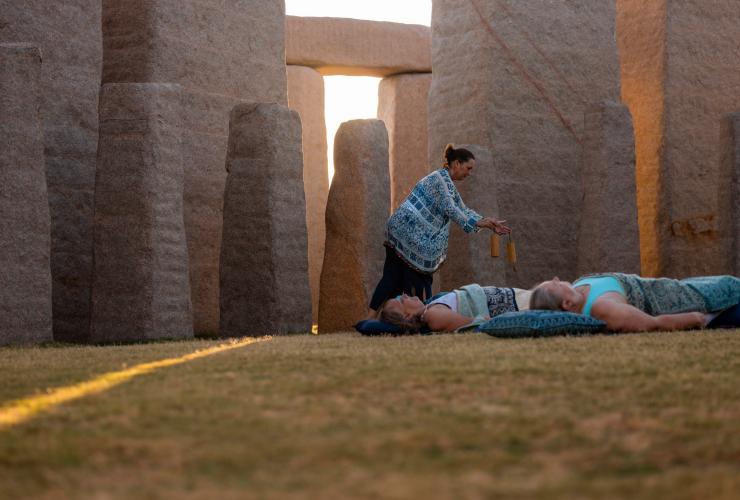 Ospiti stesi su tappetini da yoga con un cuscino sull'erba tra strutture di pietra, con un'insegnante che muove degli strumenti sopra di loro durante un ritiro con Untamed Escapes, Esperance, Western Australia © Tourism Australia