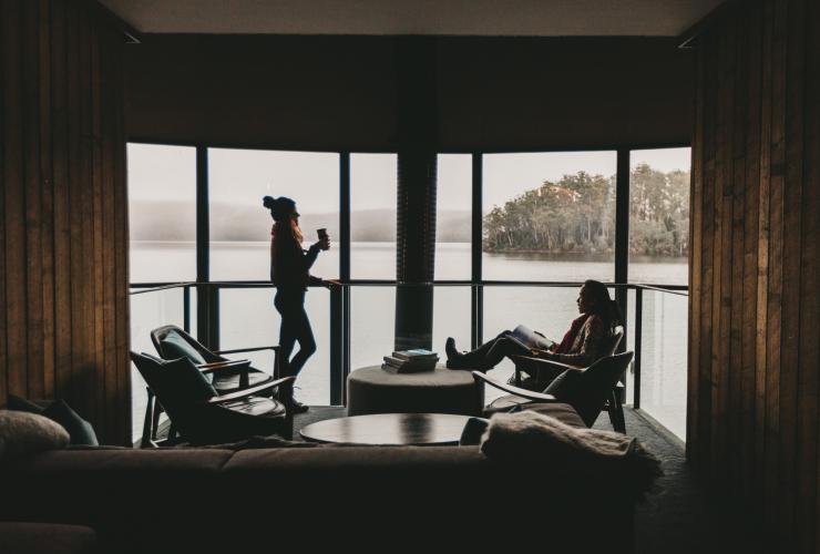 Uomo e donna che si rilassano davanti alla finestra ammirando i panorami sconfinati di Lake St Clair a Pumphouse Point, Tasmania © Jarrad Seng
