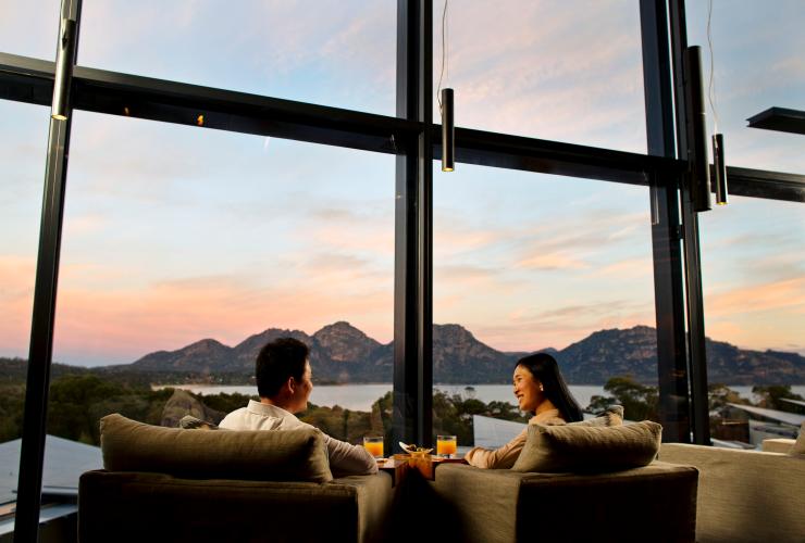 タスマニア州、サファイア・フレシネの夕暮れ時、コールズ・ベイの山と海を見下ろすラウンジに座るカップル © Myka Photography